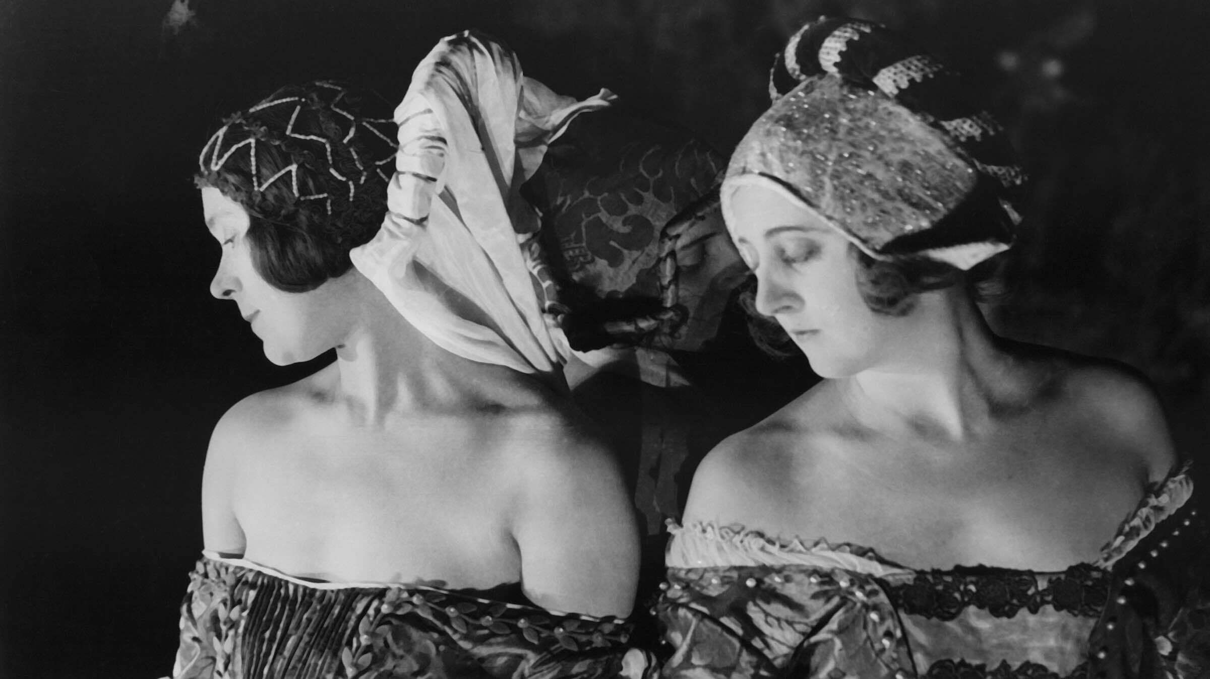 B&W photo of two women in headresses and dresses standing next to each other, looking down, somber