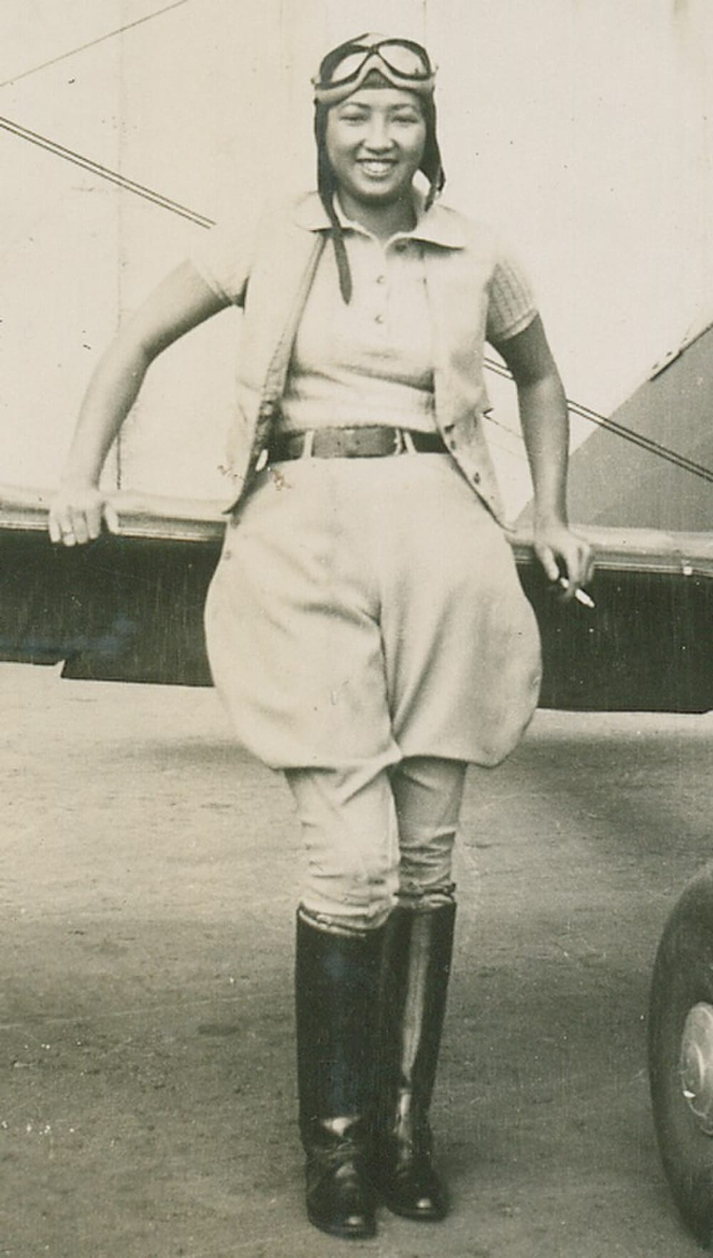 Sepia toned portrait photo of Lee in an aviator suit, stands in front of the wing of a small plane.