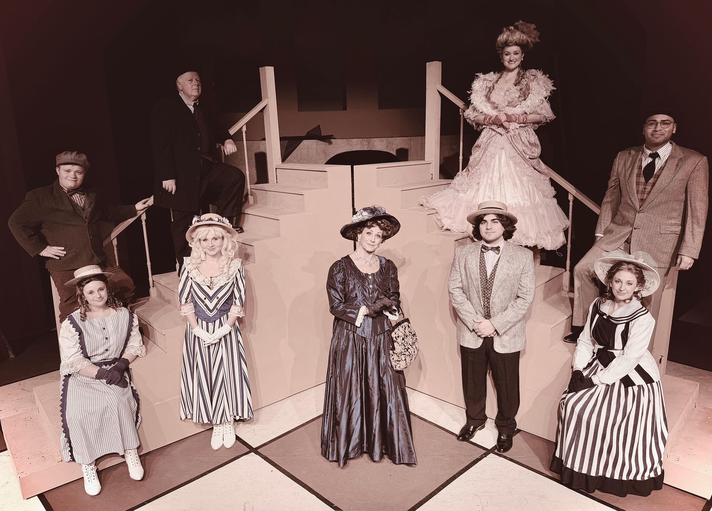 A group photo of the cast on stage, wearing early 1900s fashion suits and dresses and bonnets and top hats.