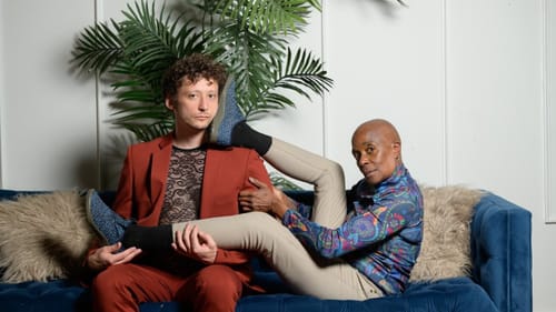 Grinberg and Moore pose together on a blue couch with a large plant behind them. Their bodies interlock in interesting ways.