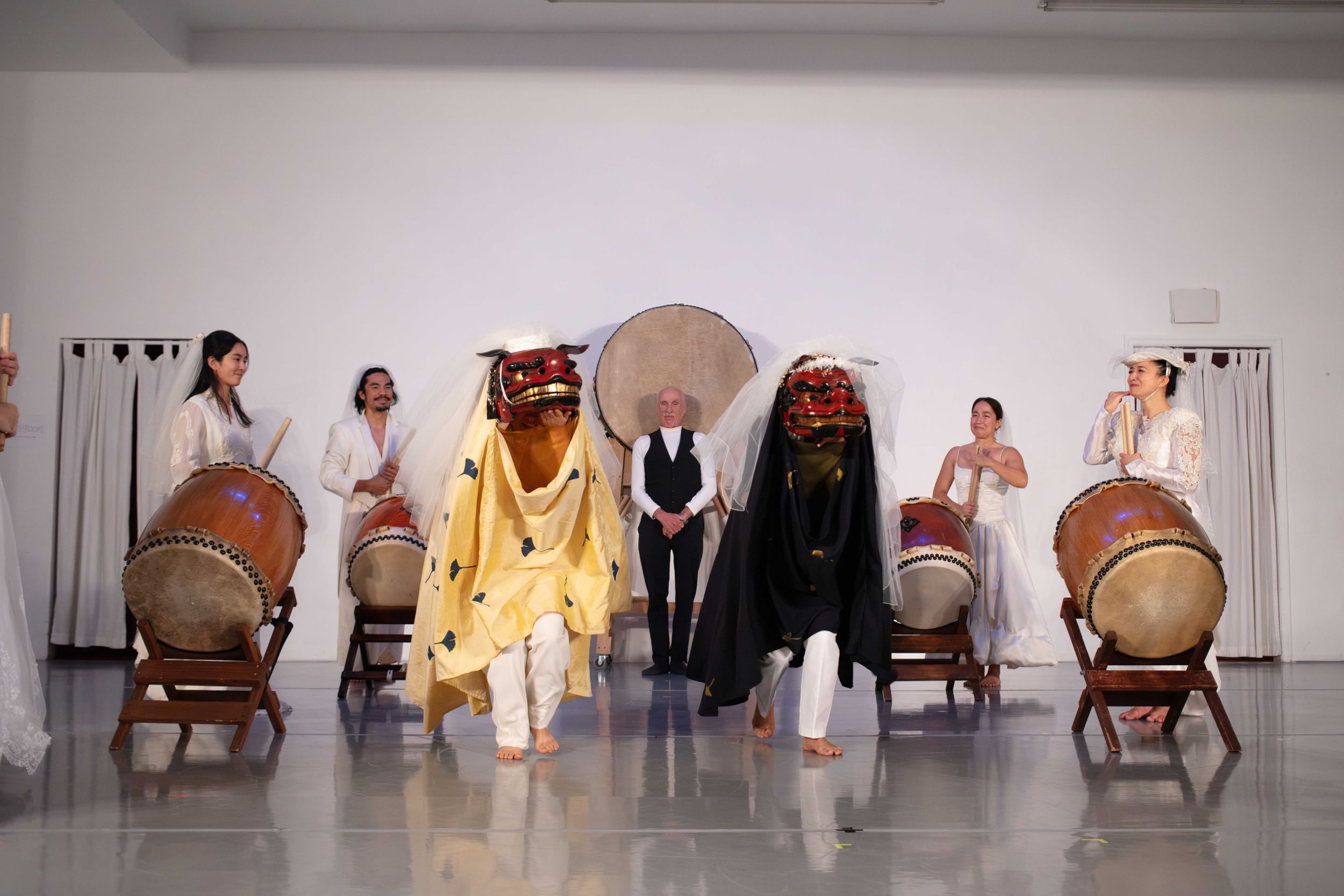 Taiko drummers in bride-like white outfits surround two dancers wearing wooden Lion Dance heads with golden teeth.