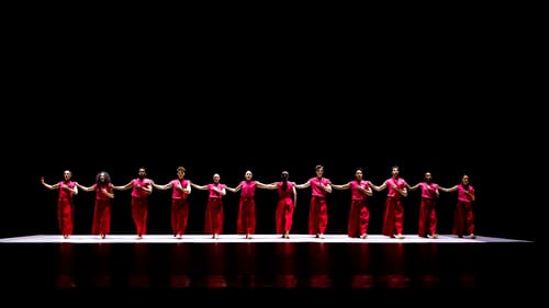 A line of 12 dancers in red costumes stand with their right arm extended to the shoulder of the next person.
