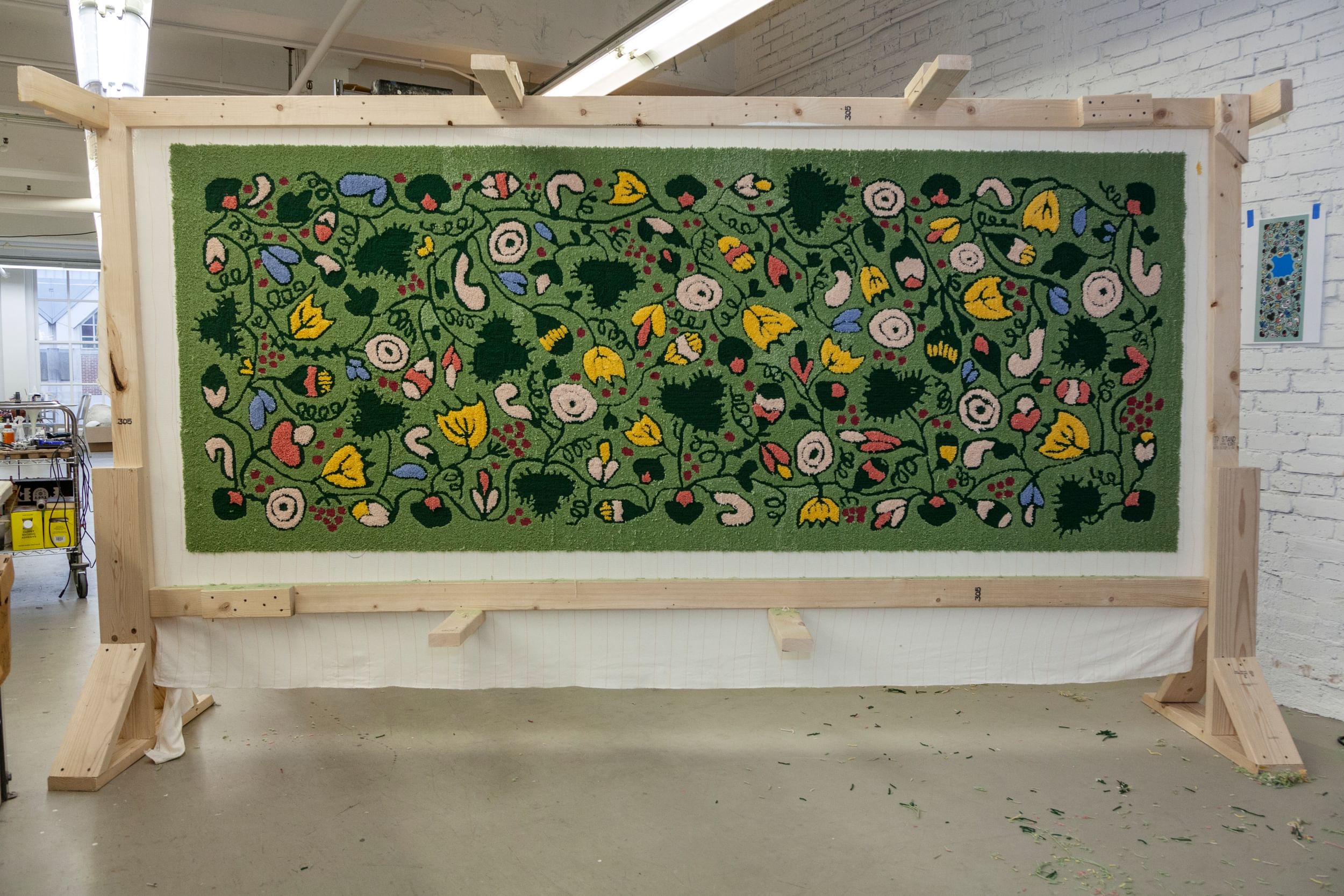 A 4x8-foot rug mounted vertically. Blue, yellow, peach & white flower designs loop on a green field.