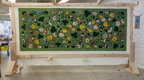 A 4x8-foot rug mounted vertically. Blue, yellow, peach & white flower designs loop on a green field.