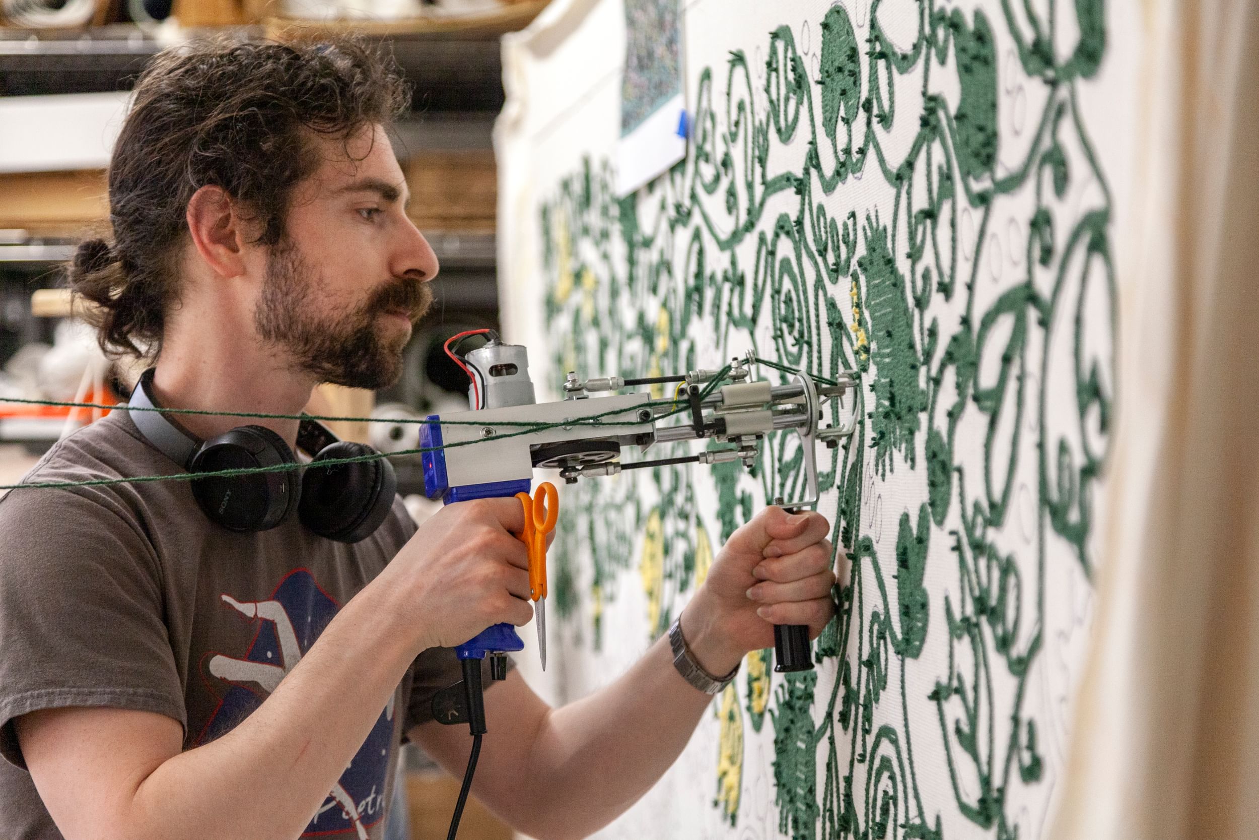 West, a bearded man in profile, uses a tufting gun to pierce a pale stretched fabric canvas with green thread.