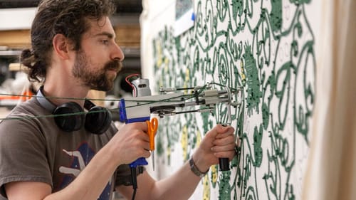 West, a bearded man in profile, uses a tufting gun to pierce a pale stretched fabric canvas with green thread.