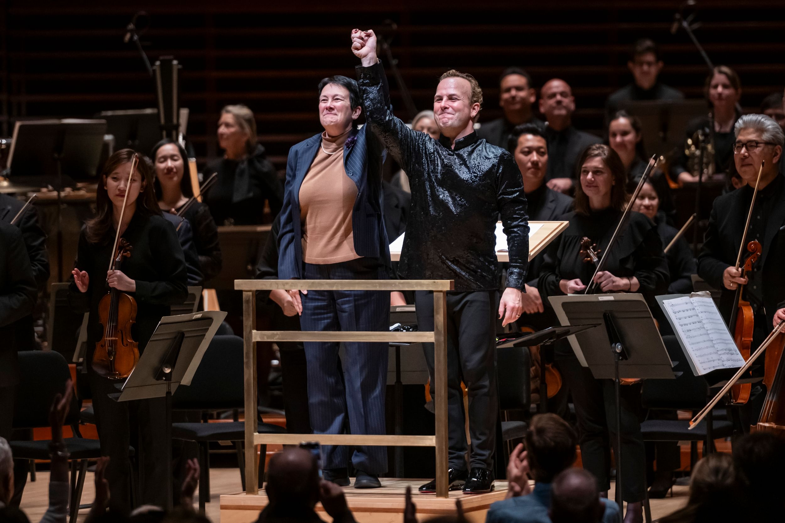 Higdon & Nézet-Séguin stand together on the podium, orchestra around them, smiling and raising their clasped hands.