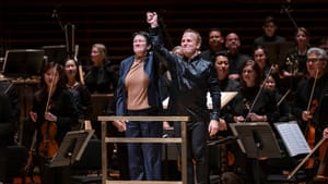 Higdon & Nézet-Séguin stand together on the podium, orchestra around them, smiling and raising their clasped hands.