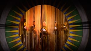 Row of actors in a patterned circular frame, obscured by a curtain of gold fibers which they thrust their hands through