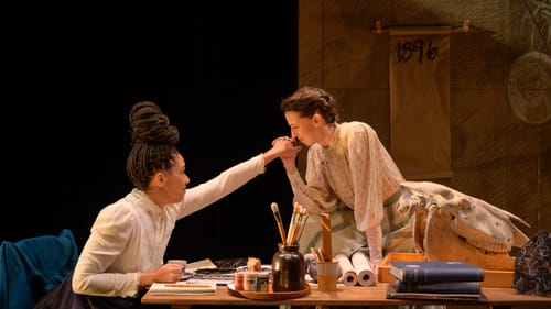 Moliére sits at a table with art supplies & books. Sieh sits on the table clasping Moliére’s hand and drawing it to her mouth