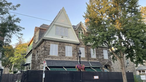 A boxy Romanesque revival square stone building with multiple roof peaks behind a black construction fence on the sidewalk.