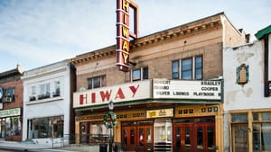 A movie theater in the middle of a small town street, shops on the block. Hiway is spelled out on the theater billboard