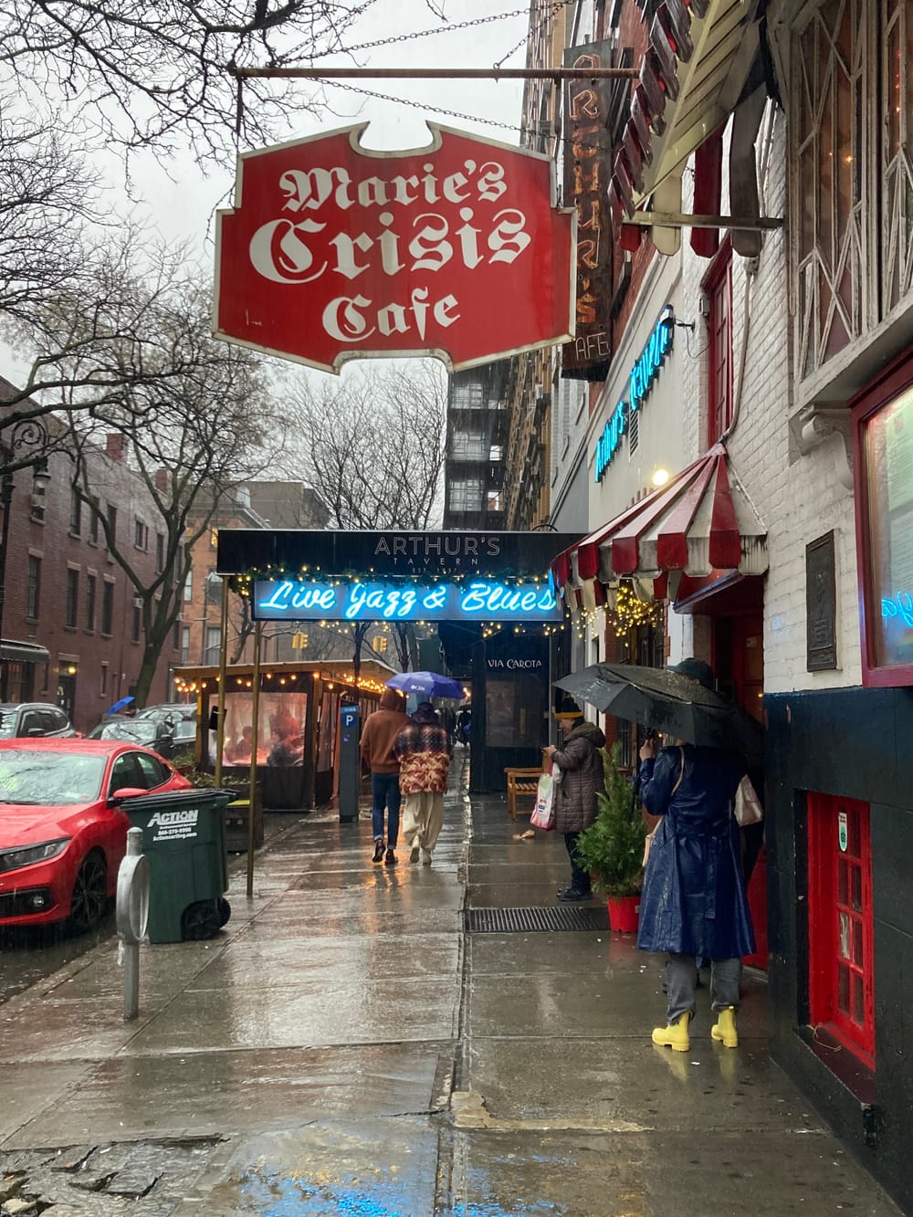 Photo of a rainy New York City street and the red hanging sign for Marie’s Crisis Café, with ornate calligraphic lettering.