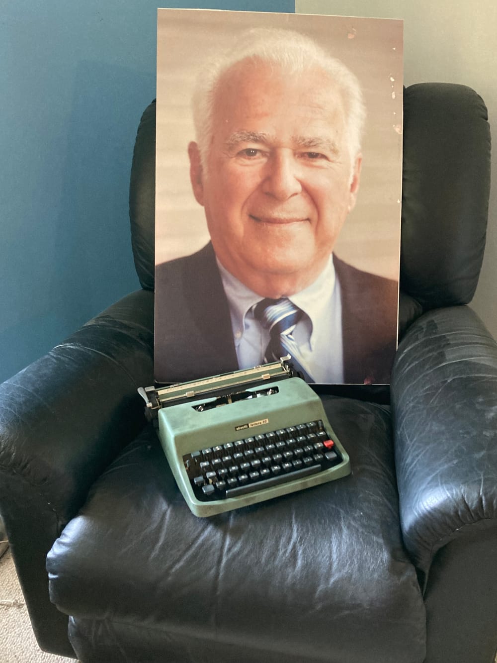 A large photo of the writer’s dad, a white man with white hair, placed on top of a worn recliner chair and vintage typewriter