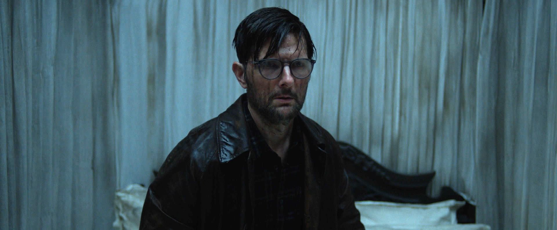 Scott, a white man with messy short dark hair, glasses, and a leather jacket, face streaked with mud, looks out with dread
