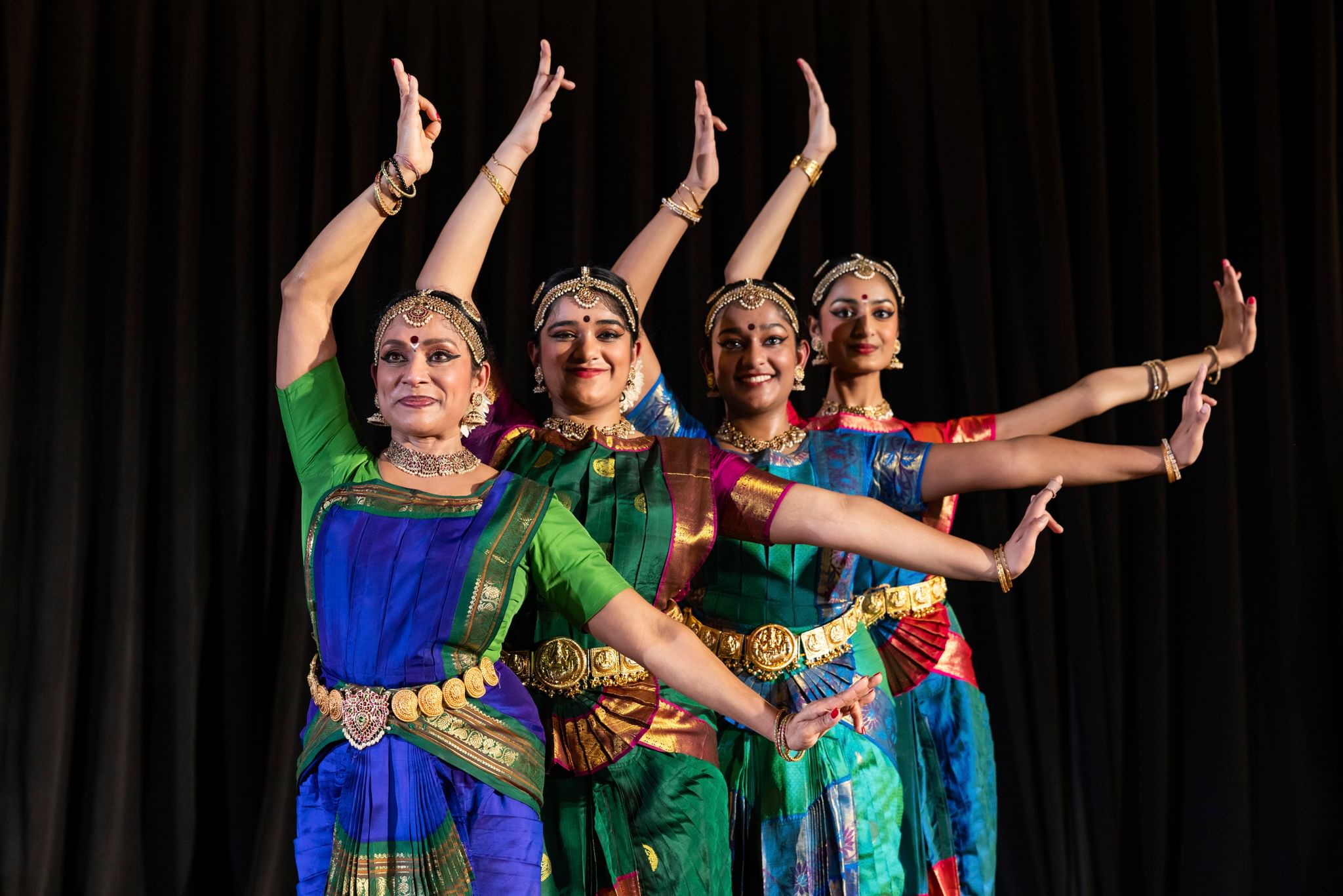 Four Indian women in colorful sarees, headwear, and bracelets on stage, all with right arm up, left extended out, in sync