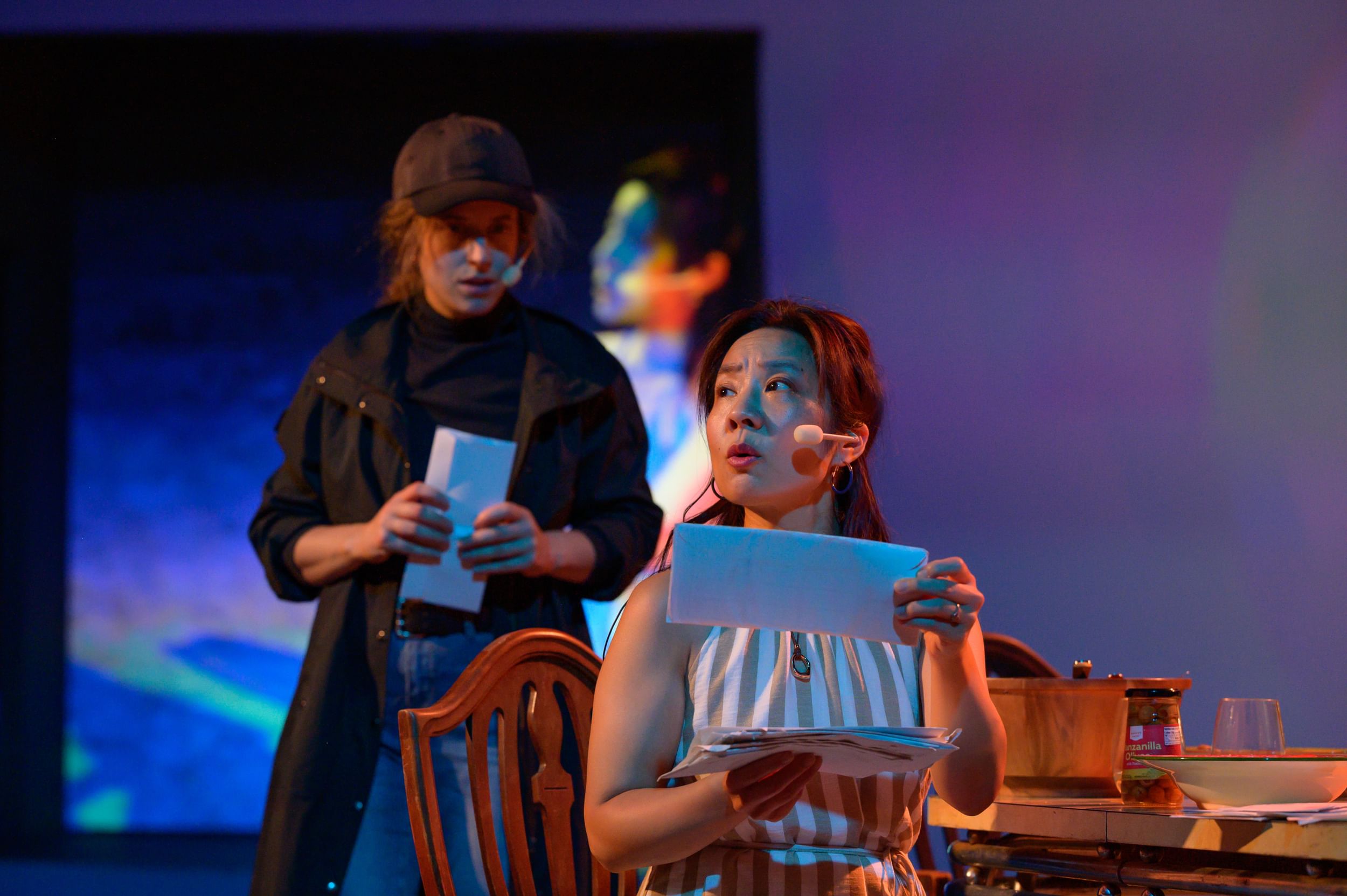 Ngo, in a striped sleeveless dress, sorts envelopes, looking concerned. Gold, in black jacket & cap, looks in intently behind