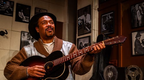 In a corner of a music studio, Beam smiles and plays a guitar. He wears a hat and a sweater with brown collar and sleeves.