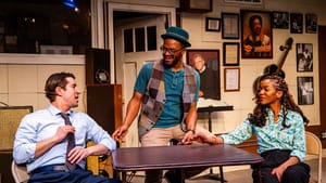 The three actors sit around a small table in a music studio full of amps, framed photos, and guitar. They talk, smiling.