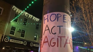 Nighttime city photo of a light pole with a handwritten sign on it that says hope, act, agitate.