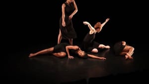 Women dancers in black pose on a black stage. Two lie on their sides; one rises w/ arms lifted, & one stands bending an arm