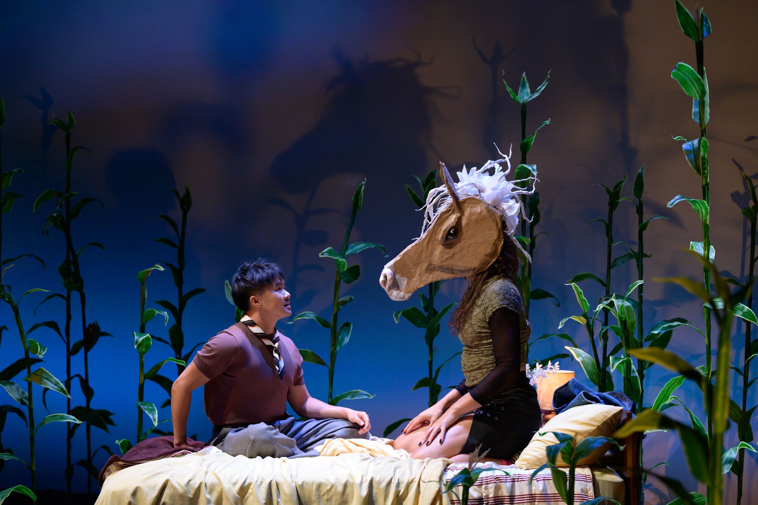 The actors sit together on a bed surrounded by cornstalks. Ni Ni looks lovingly at de la Brisa, wearing an artful horse head