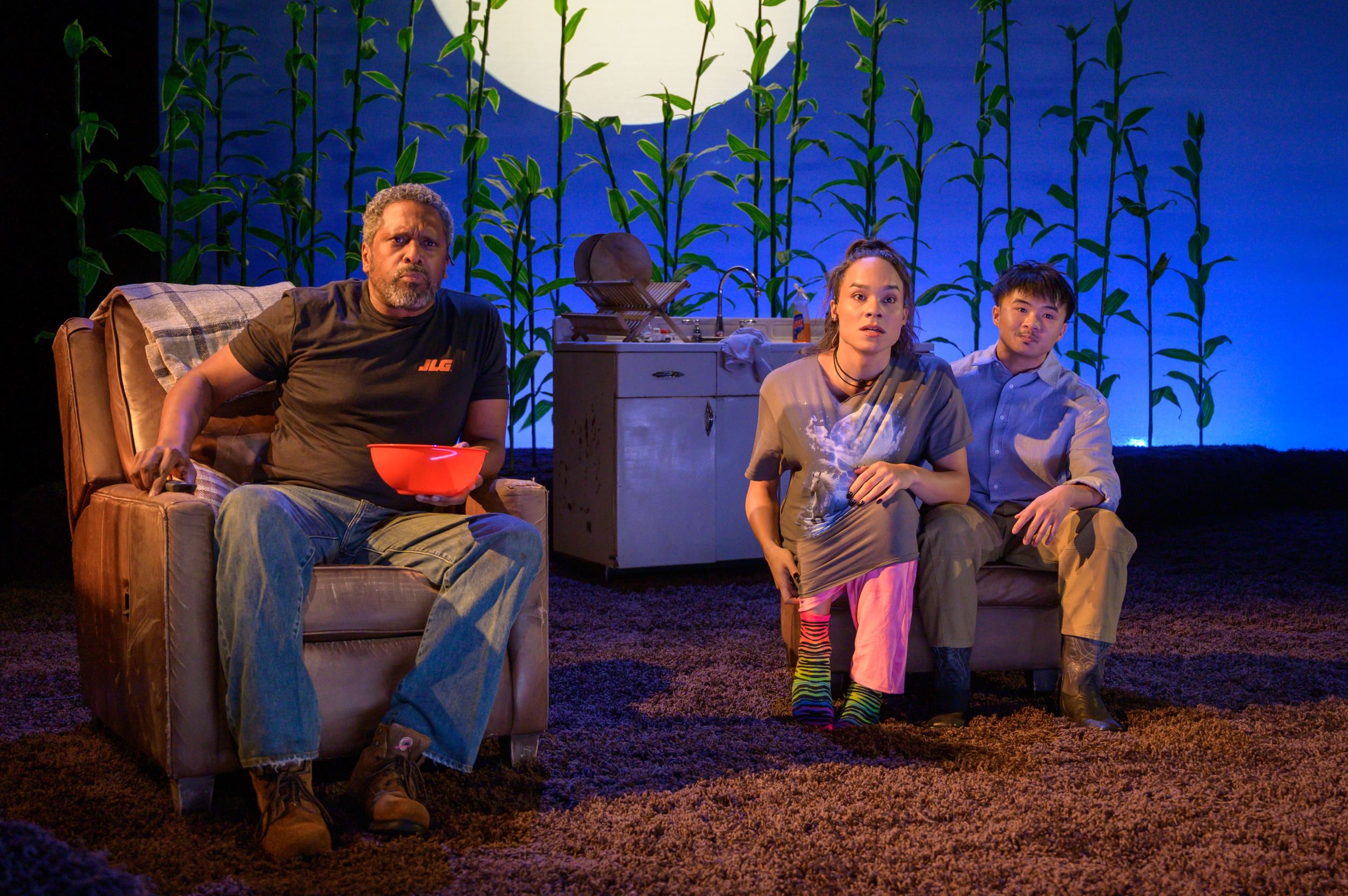 On surreal but homelike furnished stage with cornstalks behind them, the three actors face the camera, avidly watching TV