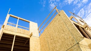 View from below the wooden framing of a house under construction, against a bright blue sky.