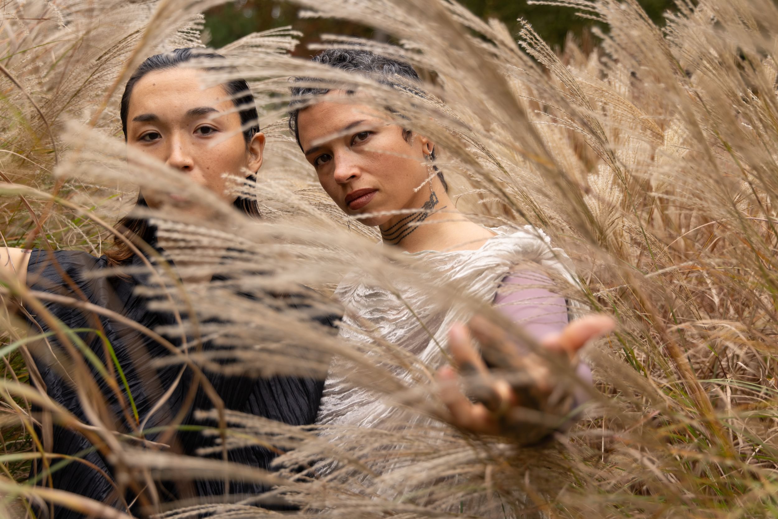 kwon, wearing black, poses with Andrews, wearing white, partially obscured by soft, tall grasses that reach over their heads.