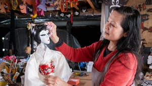 Zhang places flowers on a Geisha doll that has white, black, red paint as makeup and a ivory kimono in a studio