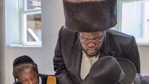Black, wearing a shtreimel, with a Black boy in Orthodox Jewish attire, leans over another Orthodox man writing on a scroll.