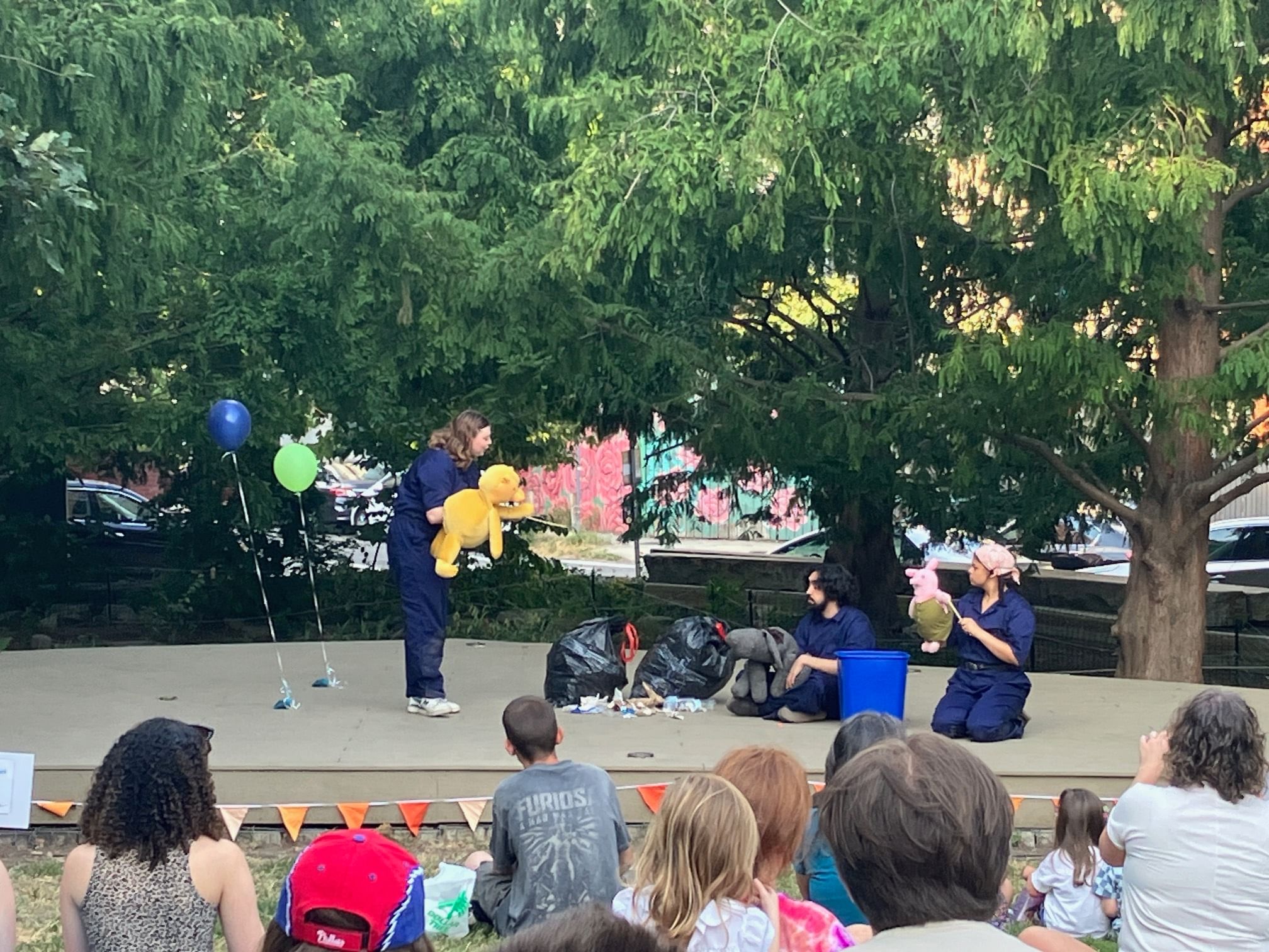Three puppeteers in all blue on stage with Winnie, Eeyore, and Piglet puppets, full trash bags, balloons, outside in a park