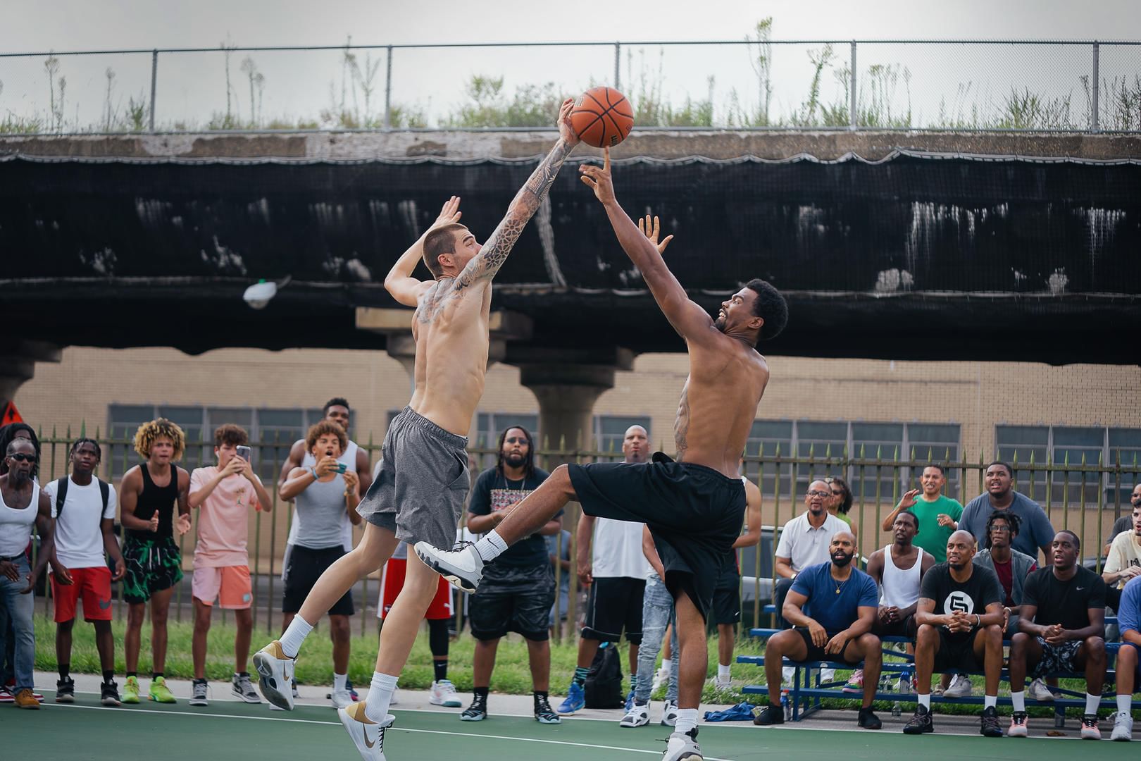 Film still: two leaping basketball players fight for the ball on an outdoor daytime Philly court in front of about 25 people