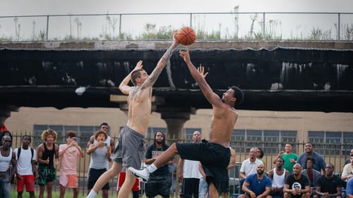 Film still: two leaping basketball players fight for the ball on an outdoor daytime Philly court in front of about 25 people