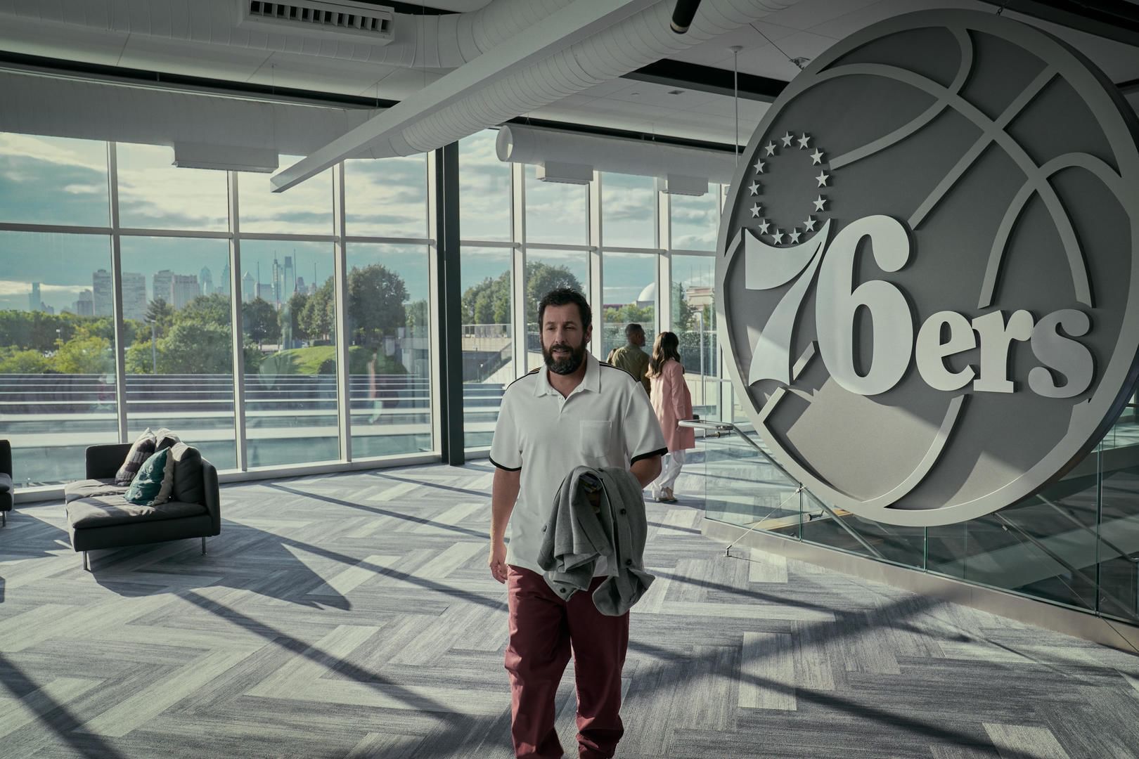 Film still: Sandler walks through the gray-carpeted Sixers lobby, the Philly skyline visible through the window behind him.