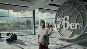 Film still: Sandler walks through the gray-carpeted Sixers lobby, the Philly skyline visible through the window behind him.