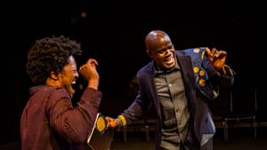 Vaughn & Thomas, Black male actors, dance together. Vaughn has an afro & red button-down; Thomas is bald & wears a blazer.