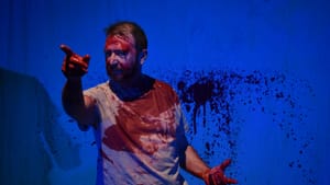 Smith, a white man in a white tee, points intensely in front of him. He’s splattered with red paint like wet blood.