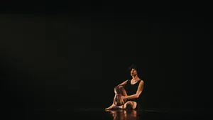 A female dancer calmly sits alone on a dark stage, the warm light on her forming a floor reflection of her body.