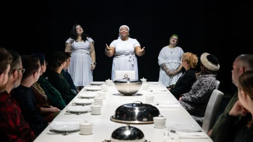 A white dining table with the audience sitting at it, and three cast members in white singing at the far end.