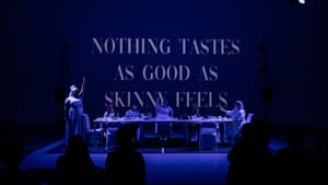 The blue-lit actors toast each other with forks. Words projected behind them say "nothing tastes as good as skinny feels"