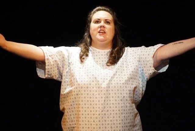 Snapp, a white woman with long brown hair, stands spreading her arms in a hospital gown.