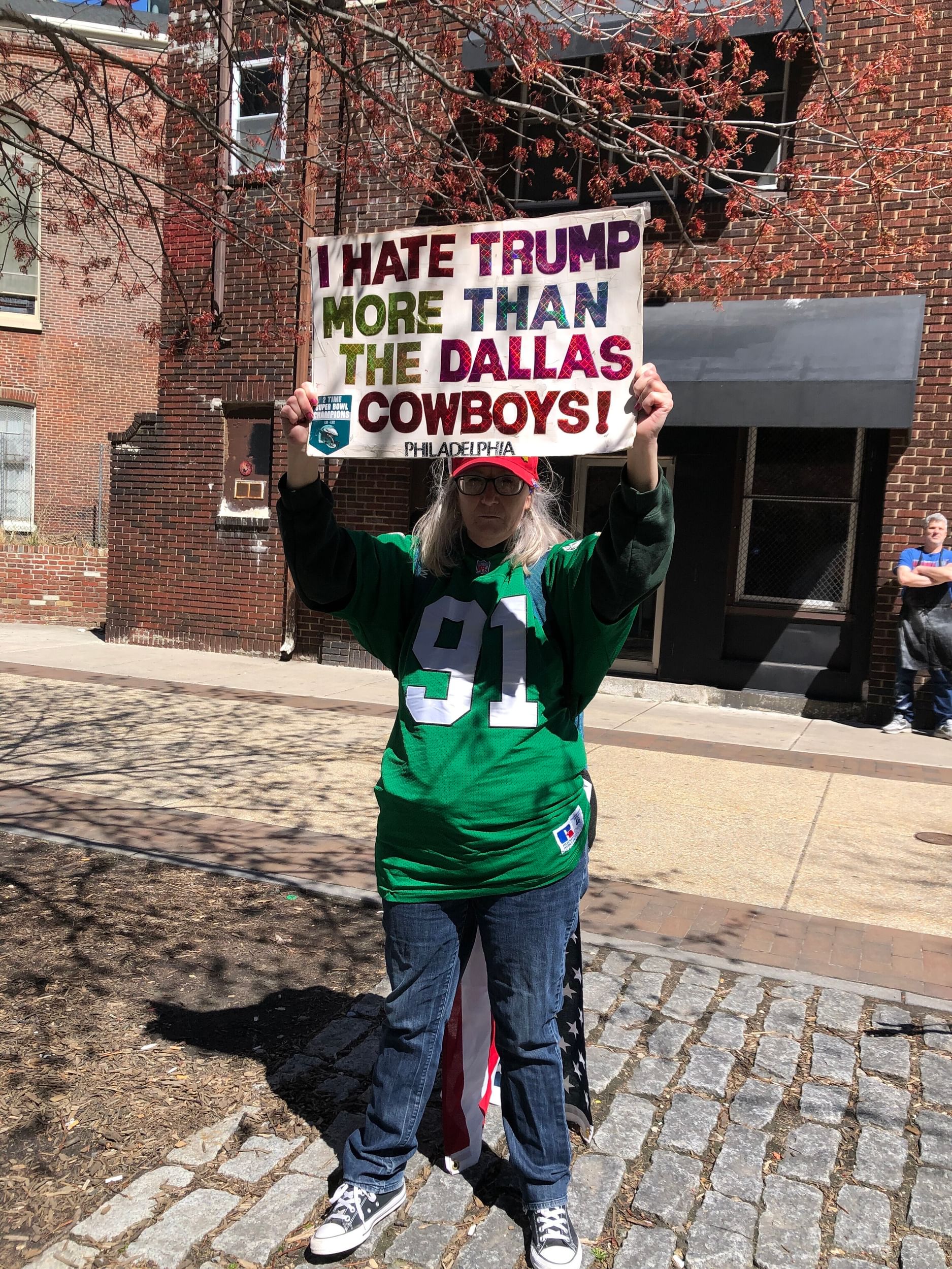A woman in a green Eagles jersey stands alone holding a sign that says “I hate Trump more than the Dallas Cowboys!”