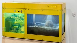 Gallery photo of Cheng’s work, a rectangular yellow box with water pouring on a rock on one side & rocks waiting on the other