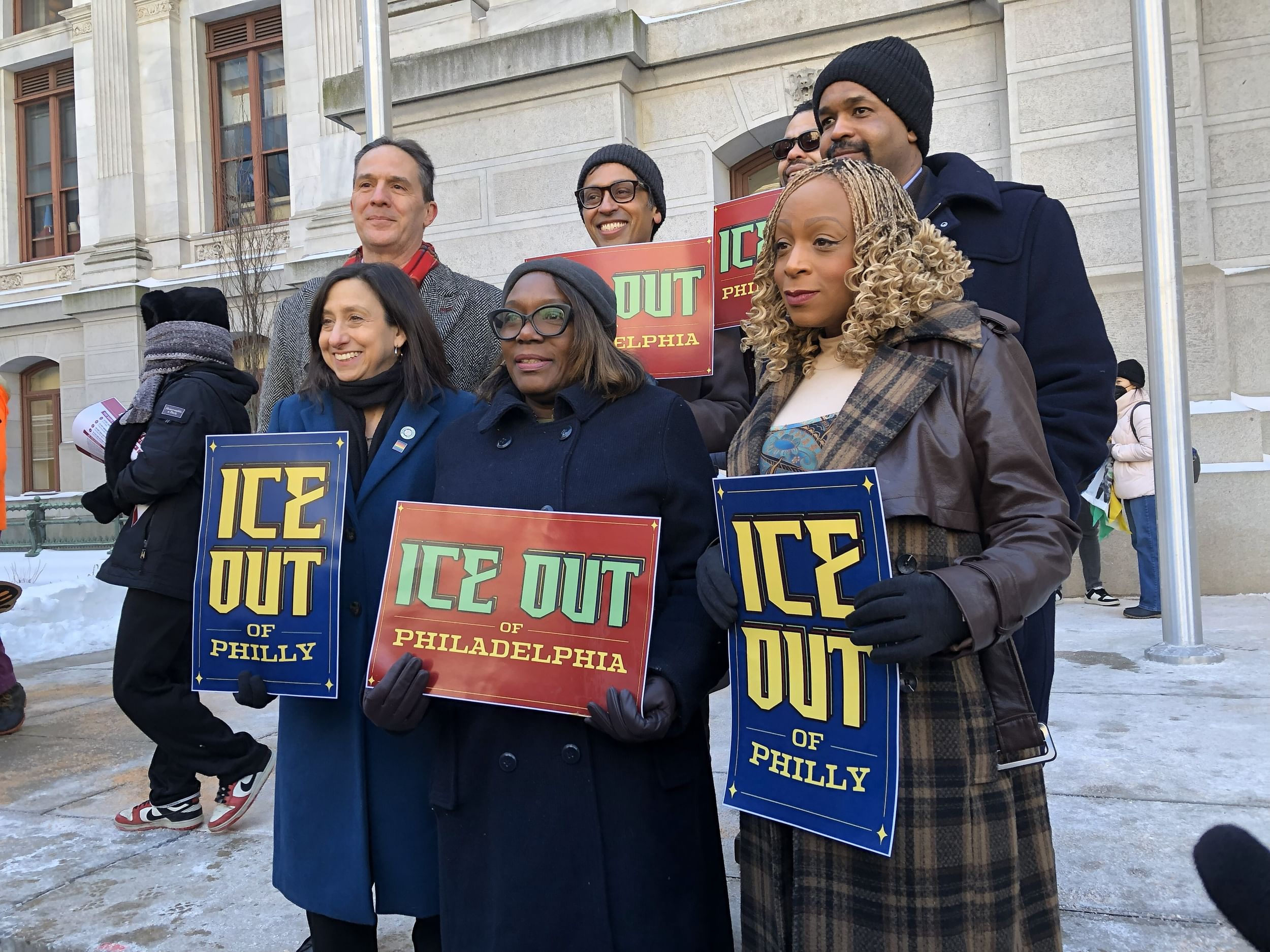 Diverse group of seven legislators pose together on a freezing day, holding signs that say “ICE OUT”