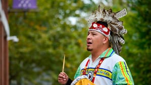 An Indigenous person dressed in colorful patterns & headdress holds a drum stick in hand, a small instrument in the other