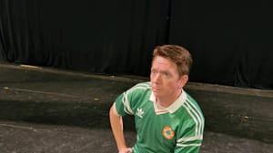 Conallen, a white man wearing jeans and green soccer jersey, sits onstage on a bench, holding some paper & looking nonplussed