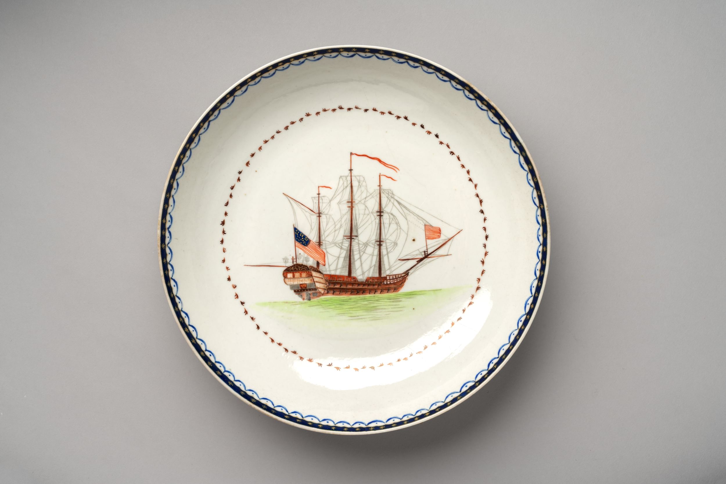 18th-century white porcelain bowl showing a 3-masted merchant ship on a green sea, with a US flag flying at its rear.