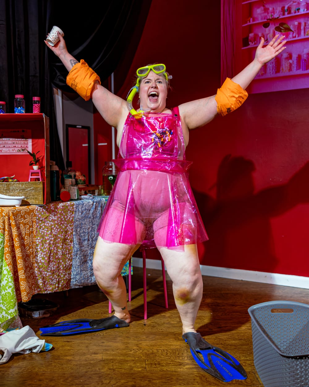 Carson dressed in pink tights, with a clear apron, flippers, and floaters on her arm. Arms out extended, exclaiming excitedly