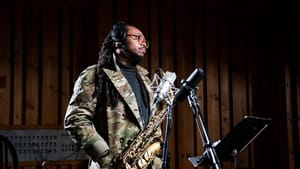 Wilkins, a Black man, in a camo-like blazer jacket holds a saxophone by a mic and conductor stand in a studio space